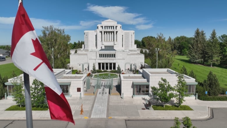 A Canadian flag flies in the foreground. An elaborate building amid greenery is seen in the background.