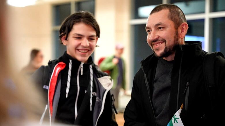 Two men, one a father and one a son, smile at someone off camera. 