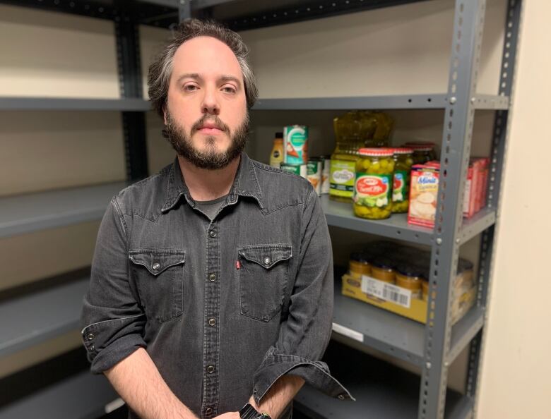 A person stands with their hands folded in front of some metal shelves. The shelves are mostly empty, with some non perishable food items on one shelf to their right.