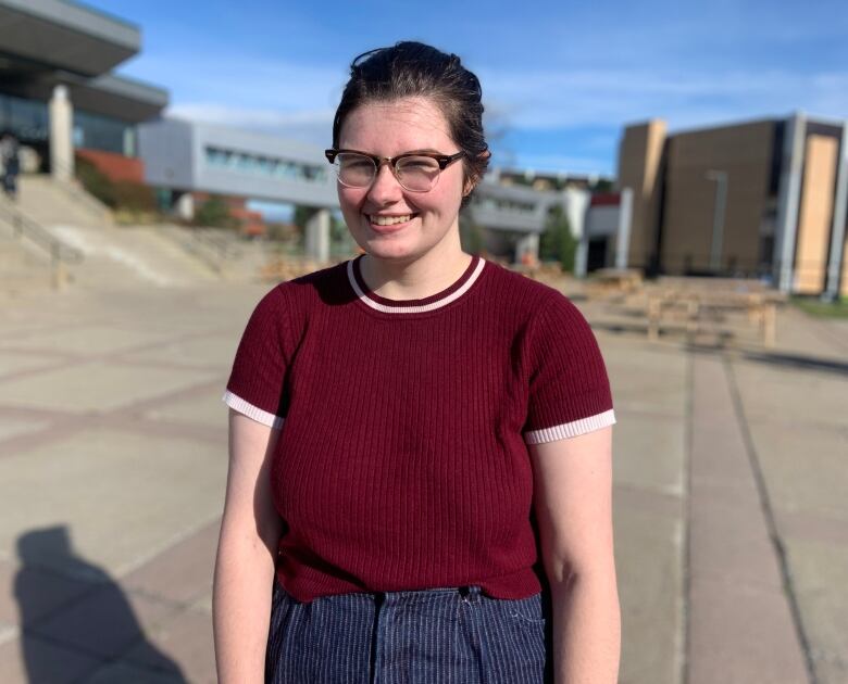 A person wearing glasses and a red t-shirt stands outside. A pedway and a blue sky can be seen in the background.