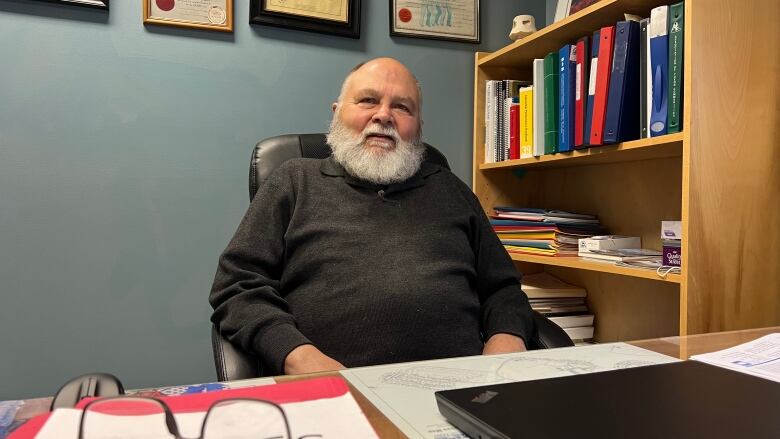 A man with a white beard sits in an office at a desk.