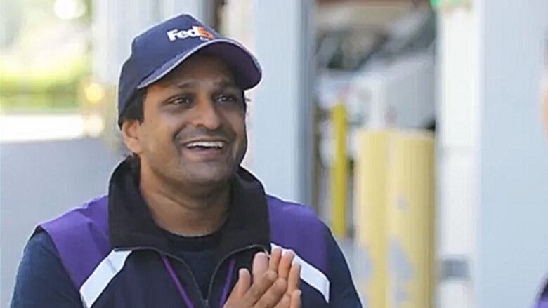 A medium shot photo of a man with his hands together in front of him and a smile on his face and a fedex hat on his head