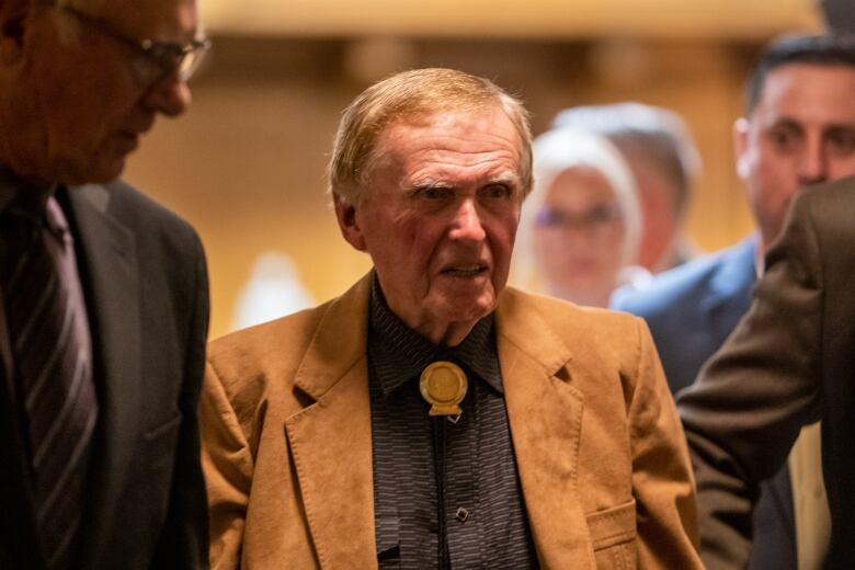 A man in a brown jacket and bolo tie stands among other people.