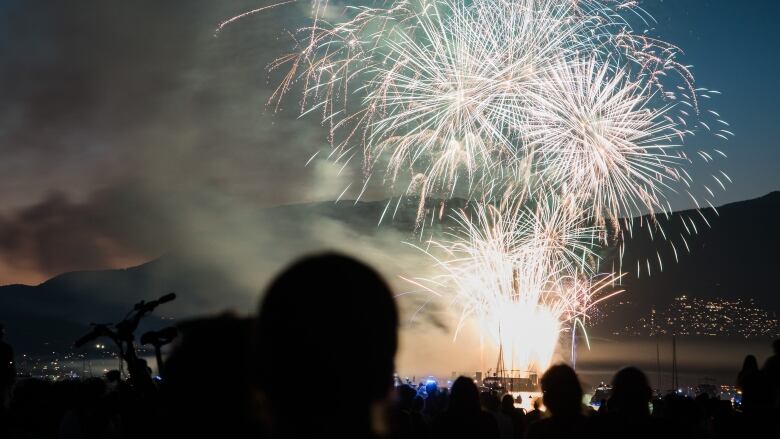 White fireworks explode in the sky above the ocean as people watch in the foreground.