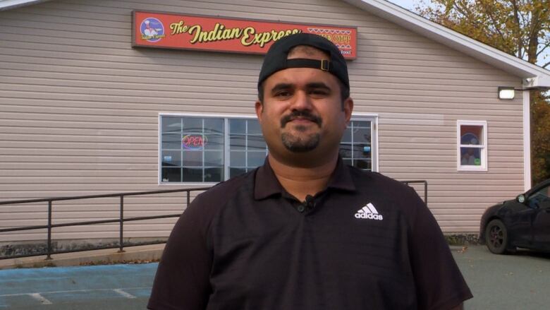 A man wearing a black shirt and baseball cap stands in a parking lot while smiling for the camera.