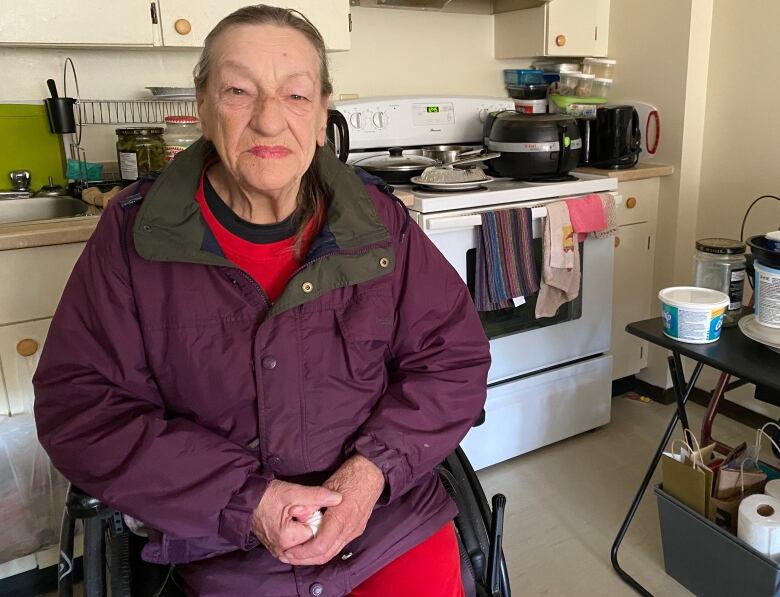 A woman wearing a purple jacket sits in her wheelchair in her kitchen. 