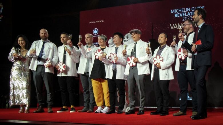 Eight people in white jackets holding glasses of champagne and a white star symbolizing their entry into the Michelin Guide for restaurants stand on a red-carpeted stage flanked by a woman in a silver dress and a man in a suit.