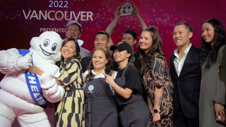 A group of nine people celebrate at the Michelin Guide awards, with one of them hugging a person dressed as the Michelin Man.