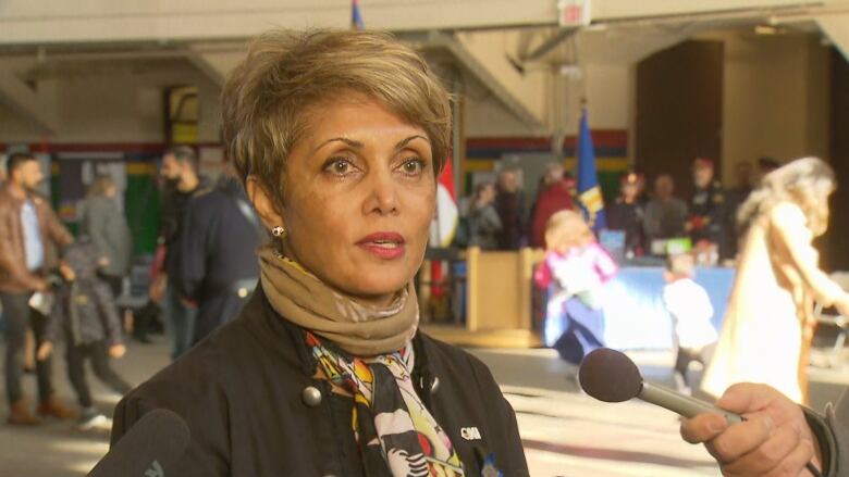 Jyoti Gondek is being interviewed by reporters, you can see one microphone to your right placed in front of her. She has short sandy blonde hair, is wearing a fashionable scarf that is brown with red and a brown jacket.