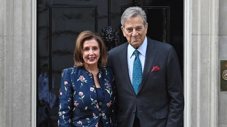 An older man and woman stand smiling side by side in front of a black door with the number 10 on it. 