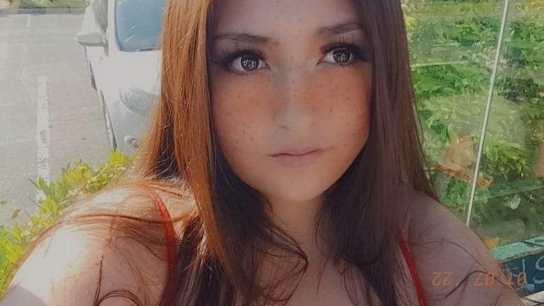 A young girl with long auburn hair, long eyelashes and a red-orange tank top takes a selfie