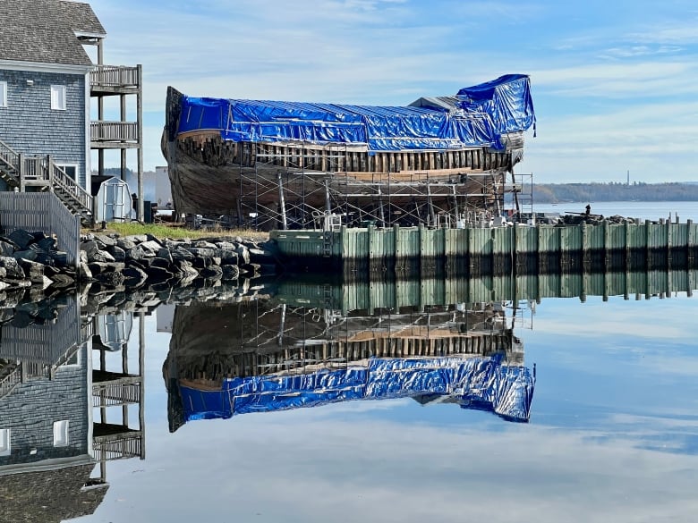 A ship undergoing restoration is reflected in still water alongside a dock.