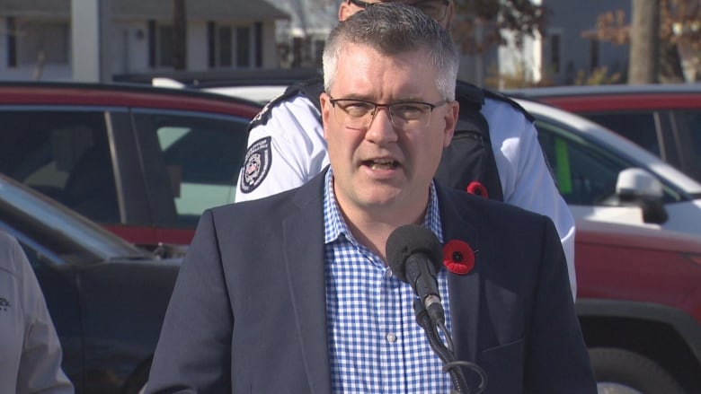 Man stands in front of cars and an RCMP officer. He speaks into a microphone.