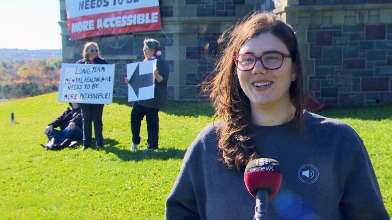 A woman with long brown hair wearing purple glasses smiles in front of a microphone. Two women stand behind her with posters, one reading 'Long term mental healthcare needs to be more accessible.