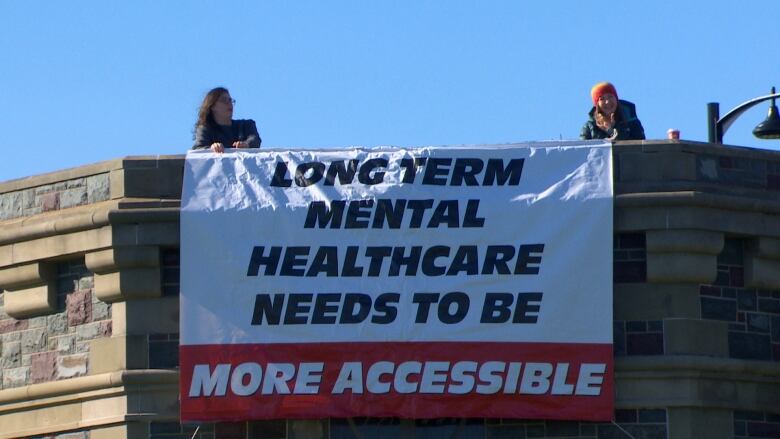 Two women stand on either side of a large banner. It's white with a red stripe across the bottom, and has black font that reads 'Long term mental healthcare needs to be more accessible.