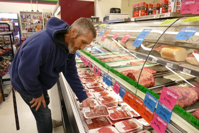 A man with greying hair and a greying beard in a blue sweatshirt looks at meat in a grocery store cabinet.