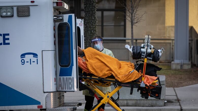 A patient on a stretcher, covered in an orange blanket, is loaded into the back of an ambulance by a paramedic wearing a medical gown, medical mask and face shield.