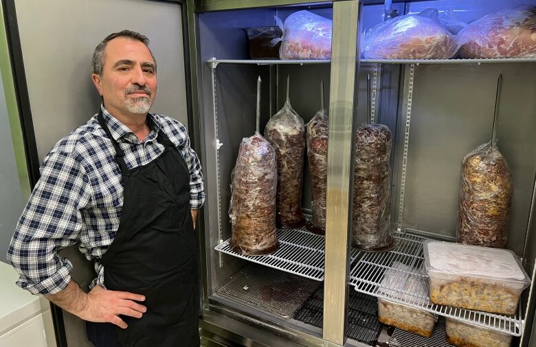 A man in a black apron stands in front of an open fridge containing upright spits stacked with slices of meat.