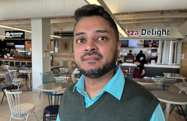 A man wearing a grey vest and green shirt standing in a cafeteria. Tables and chairs are in the background. 