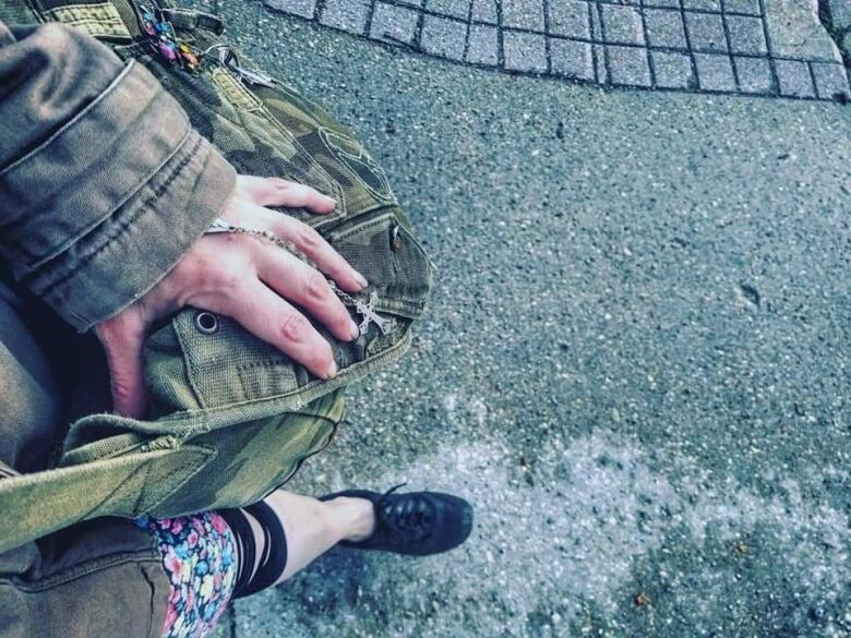 Close-up of a woman's reddened hand covering a worn-out faded green camouflage patterned tote bag, while sitting on an asphalt street.