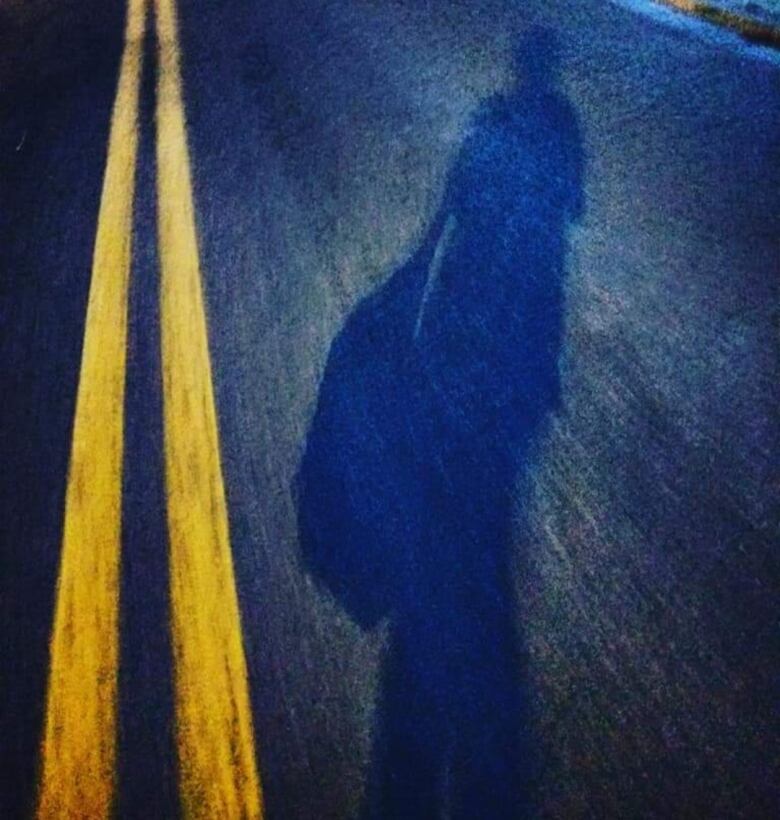 Close up of a paved street at dark, and the dark reflection of a woman walking away from the camera to the right of two bright yellow centre lines.