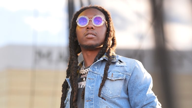 A young man with long black dreadlocks, a frayed denim jacket, and sparkly, round purple sunglasses stands outside, looking toward the sky. 