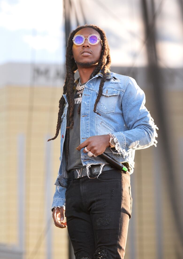 A young man with long black dreadlocks, a frayed denim jacket, and sparkly, round purple sunglasses stands outside, looking toward the sky. 