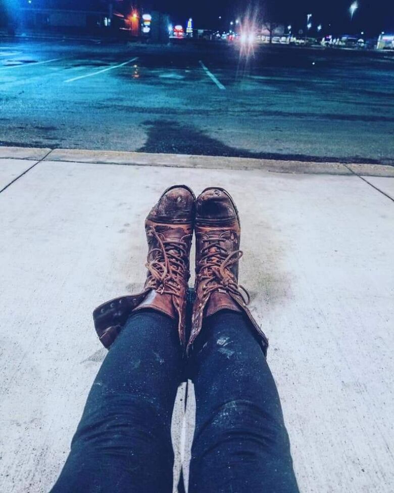 Close up of a pair of legs in blue, stained leggings and tattered brown boots, stretched across a sidewalk.