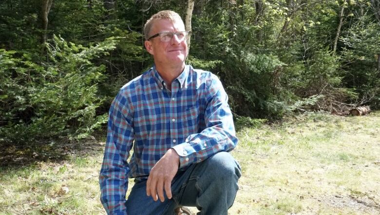 A white man in a blue-checked plain shirt and glasses kneels in the grass in front of a forest of green fir trees