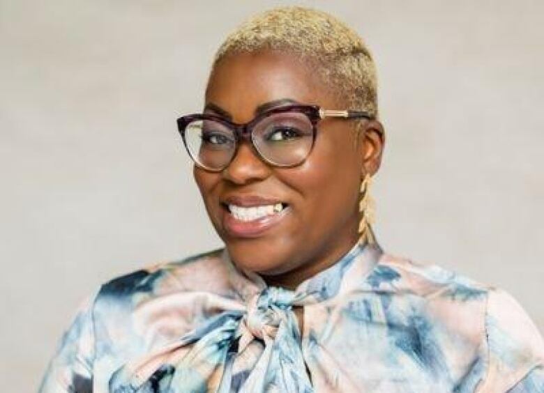 A headshot shows a smiling Black woman with close-cropped blonde hair, cat-eyed tortoiseshell glasses and dangly gold earrings.