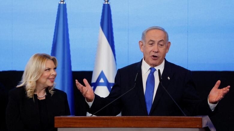 A grey-haired man standing at a podium with a flag of Israel behind him. 