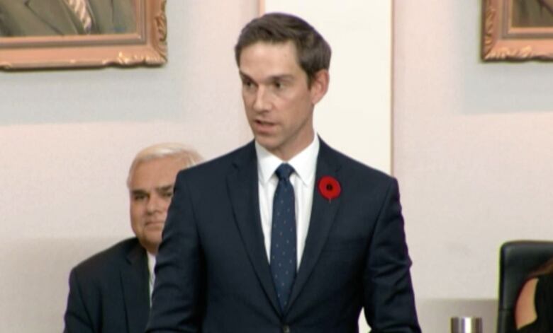 A man in a suit wearing a poppy gives a speech.