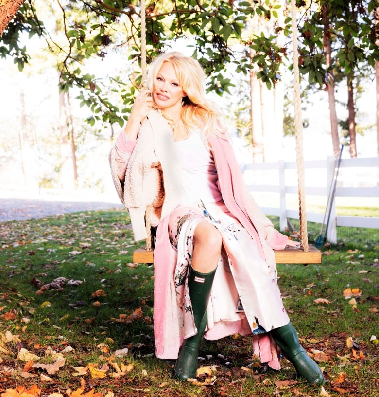 Pamela Anderson sits on a swing underneath trees covered in pink flowers. She has long blonde hair and wears a pink dress and green rain boots.