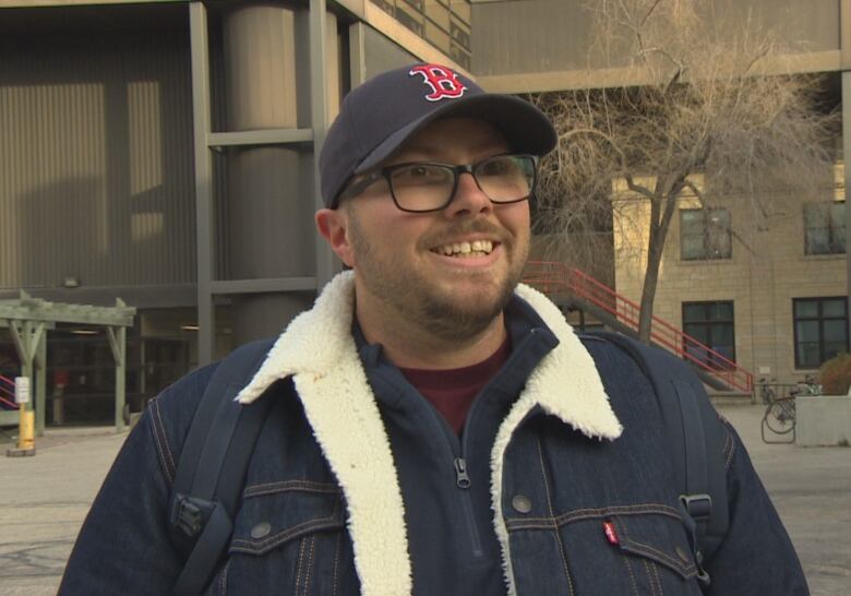 Person standing outside in jacket, ballcap, smiling.