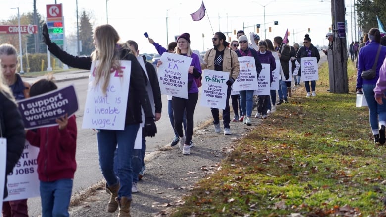 Striking workers hold signs while walking in a line on a fall day.