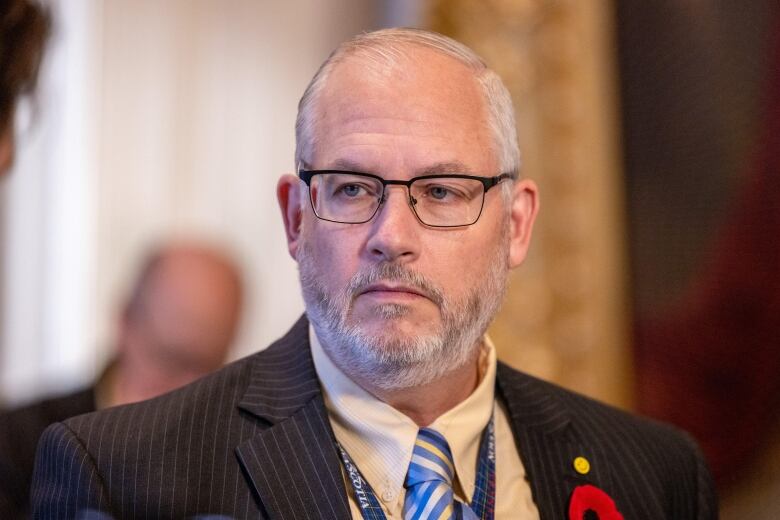 A white man with white hair wearing a suit jacket and glasses looks off to the side of the camera