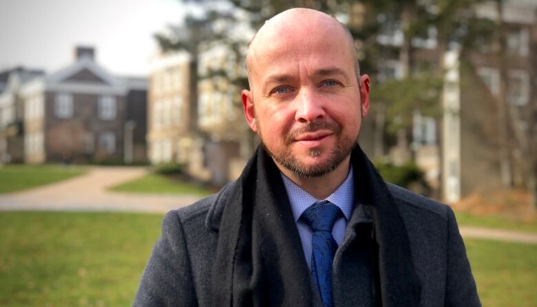 A man in a blue shirt and tie under a dark wool coat and scarf is seen standing outside, with university campus buildings in soft focus behind him. 