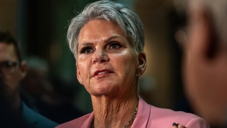 A woman with shorter grey and white hair stands in a rotunda of a legislative building wearing a pink blazer and a small microphone pinned to her clothing.