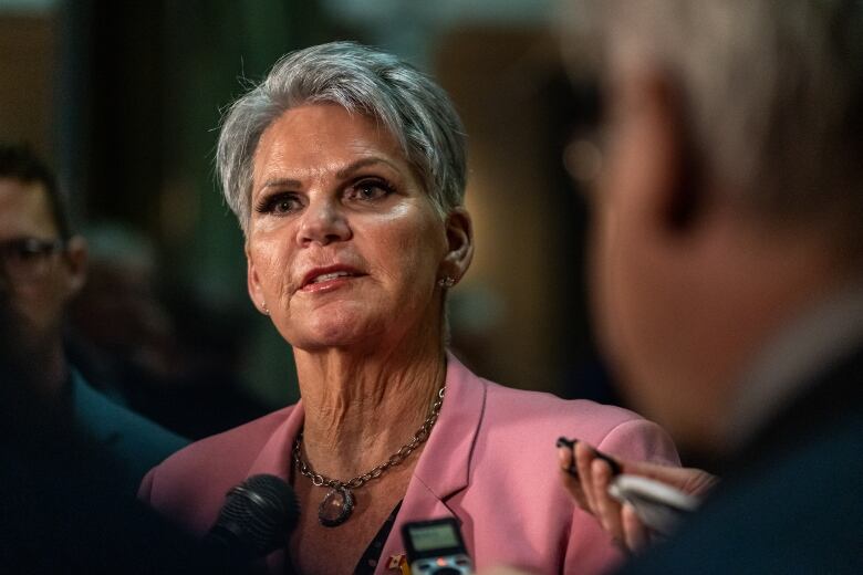 A woman with shorter grey and white hair stands in a rotunda of a legislative building wearing a pink blazer and a small microphone pinned to her clothing.