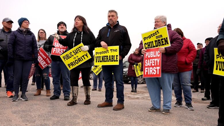 A woman speaks while holding a sign saying 'Public safety is a priority' while others look at her and also hold signs saying 'protect the vulnerable' and 'protect our children.' 