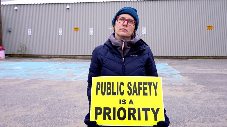 A woman in winter gear looks at the camera while holding a sign that reads 'Public Safety is a priority.' 