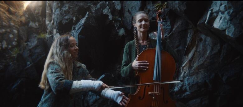 A woman holds a cello, her eyes closed. Another woman runs the bow over its strings.