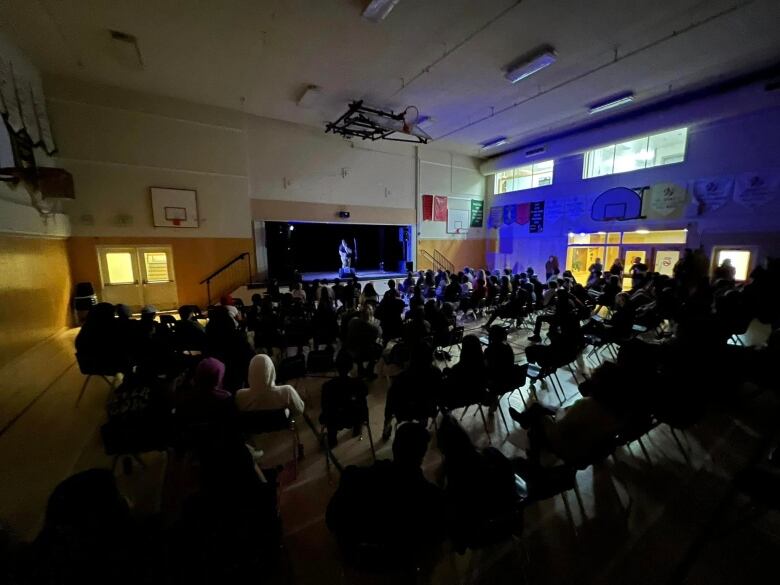 A crowded dark gymnasium as seen from the back, with a man performing on stage at the front.