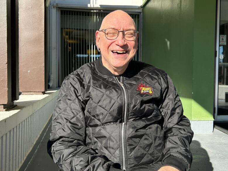 A man with a bald head and multiple piercings in his ears, wearing a black jacket with a logo that says 