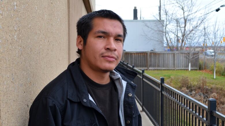 A young man stands against a wall, looking at the camera.