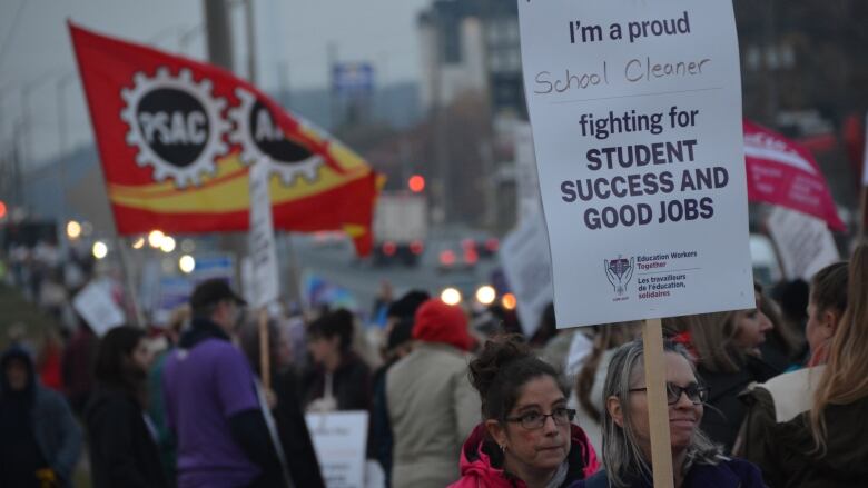 A woman holds a sign reading 'I'm a proud school cleaner' with dozens of other protesters waving flags and holding signs behind her along a busy roadway. 