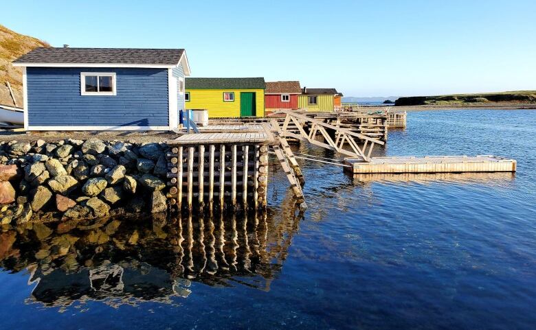 A series of fishing buildings sit atop wooden wharves by a habour. The sky and water are bright blue. 