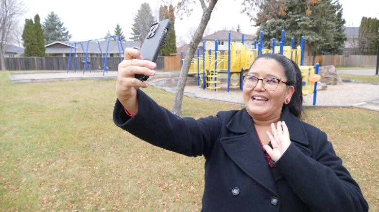 A woman wearing glasses with long black hair in a ponytail holds her phone up to take a selfie. She's waving at the camera and standing in front of a playground.