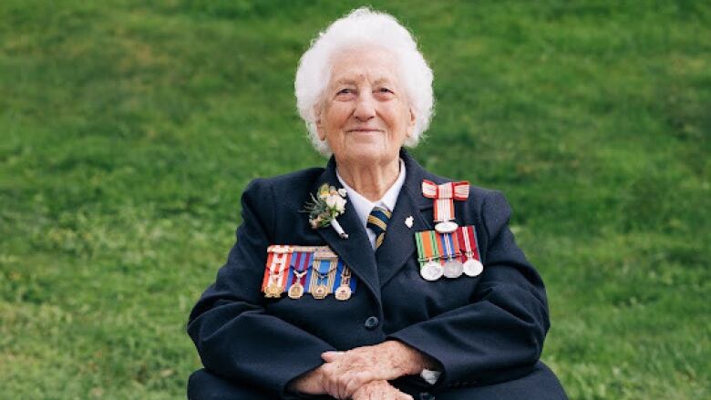 100-year-old Joan Robertson smiles at the camera wearing a jacket with her medals from the Second World War. 