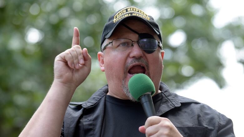 Man wearing a black cap, eye patch and black tee shirt speaks into a microphone